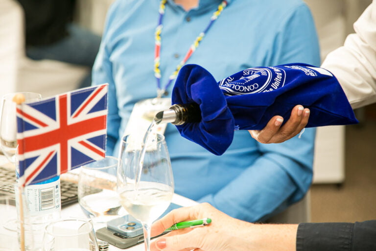 Someone serving white wine in a wine glass. The bottle is hidden in a blue CMB branded fabrik bag. There is an England flag on the table.